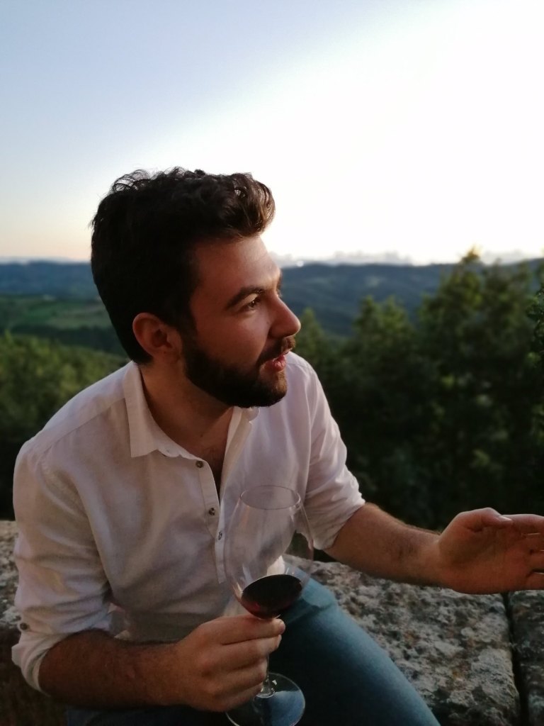 Riccardo sitting on a stone wall with a glass of red wine, green Umbrian hills behind him